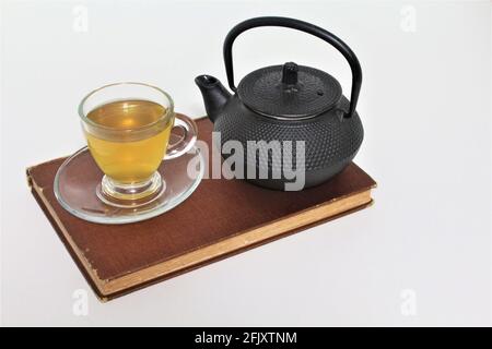Iron Japanese teapot and a cup of herbal teal in a glass teacup served on a saucer plate sitting on top of an old vintage book. Isolated in a plain wh Stock Photo