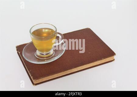 Cup of herbal teal in a glass teacup served over a saucer plate on top of an old vintage book. On a plain white background Stock Photo