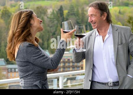 Happy laughing couple celebrating with a glass of red wine as they toast each other outdoors on the balcony of their house against a rural backdrop Stock Photo