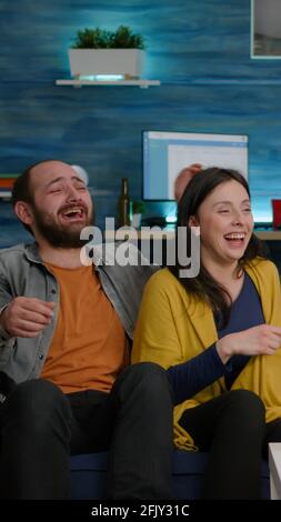 Multiracial friends laughing smiling having fun together while sitting on couch in front of TV watching comedy movie. Group of mixed race people enjoying free time drinking beer eating snacks Stock Photo