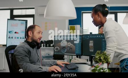 Mechanical engineer working on computer designing in CAD software 3D model of engine while african coworker asking help. Man using pc with two monitors innovating motor prototype in creative agency Stock Photo