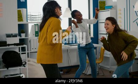 Mixed raced happy women dancing cheerfully in startup creative agency office, during work hours, enjoying victory of construction project. Afterparty in workplace, colleagues having fun in free time Stock Photo
