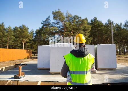The builder  talking on the phone. Architect in a protective yellow helmet and a signal vest stands backs in front of the construction site of a house Stock Photo