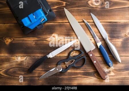 Sharpening a knife on an electric sharpener at home. Layout of knives and scissors with a sharpening machine on a wooden table. Top view, flat lay. Stock Photo