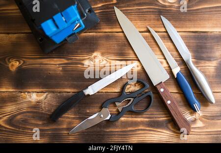 Sharpening a knife on an electric sharpener at home. Layout of knives and scissors with a sharpening machine on a wooden table. Top view, flat lay. Stock Photo