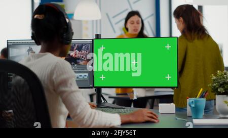 Black woman videographer employee with headphones using computer with green screen, chroma key mockup isolated display sitting in video production studio. African editor processing film montage Stock Photo