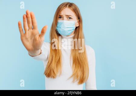 Portrait of serious young woman in medical mask standing with outstretched hand showing stop gesture over isolated blue background Stock Photo
