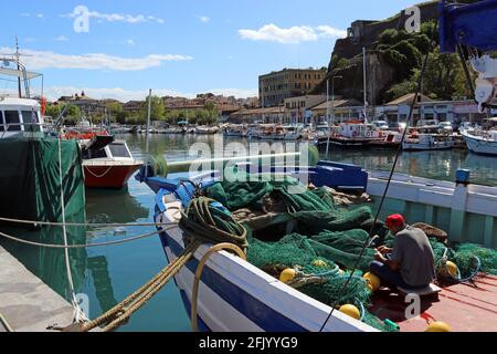 A fisherman mending nets on his boat in Corfu Town Old Harbour, below a wall of the New Fortress, right of frame.  Looking towards Corfu Old Town Stock Photo