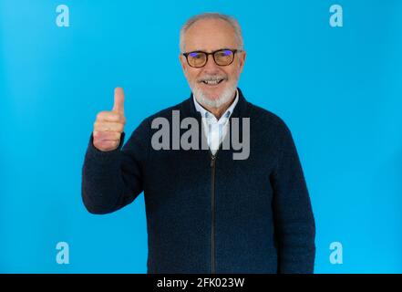 Senior grey-haired man standing over isolated blue background doing happy thumb up gesture with hand. Approving expression looking at the camera showi Stock Photo