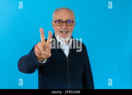 Senior grey-haired man wearing casual sweater standing over isolated blue background looking at the camera doing victory sign with fingers. Number two Stock Photo