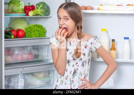 Beautiful young happy teen girl holding fresh red apple while standing near open fridge in kitchen at home Stock Photo