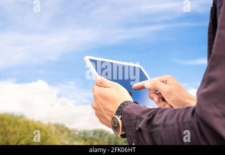 Tablet in nature outdoors. Farm or garden with blue sky and clouds. Man using smart device outside. Green grass field or park in the background. Stock Photo
