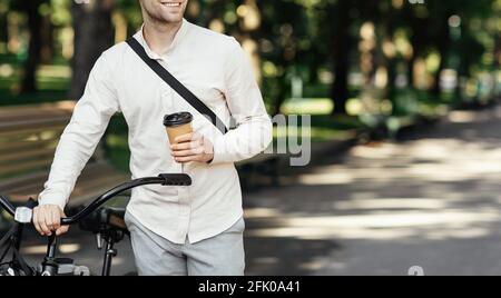 COVID-19 epidemic, morning takeaway coffee and employee on way to office. Smiling millennial man in formal clothes walks with bicycle and holds cup of Stock Photo