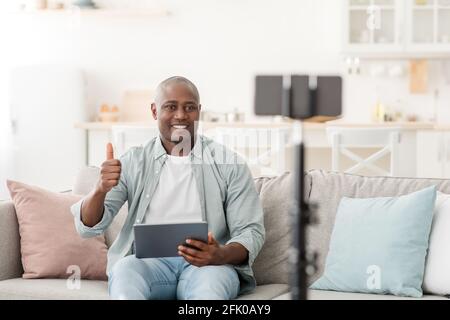 Review in video blog. African american man recommending new digital tablet, showing thumb up while recording vlog to internet channel, looking at smar Stock Photo