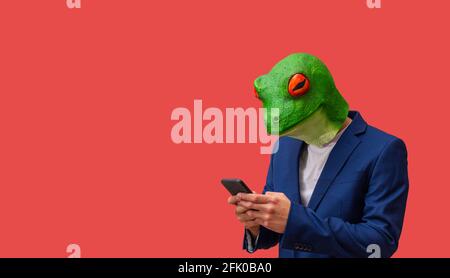 man with googly-eyed frog mask texting or watching news with smart cell phone on red background with copy space Stock Photo