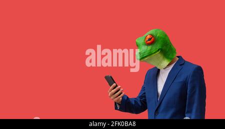 man with googly-eyed frog mask looking at smart phone on red background with copy space Stock Photo