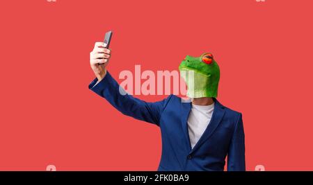 man with googly-eyed frog mask taking a selfie with smart phone on red background with copy space Stock Photo