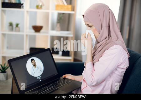 Young sick Arabian woman in hijab, suffering from cold and running nose, sits on couch at home and using laptop for video call with doctor. Female patient calling physician online. Stock Photo
