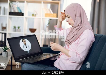 Stay at home concept. Covid prevention. Ill Muslim woman sitting on sofa at home, feeling bad and having fever, holding thermometer and calling to doctor using laptop. Online consultation with doctor Stock Photo