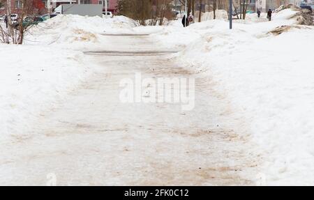 Winter sidewalk in the city, soft snow covers the path going into the distance. On the road you can see the thawing dirty snow. In the background, in the distance, houses, people, trees, a car. Stock Photo