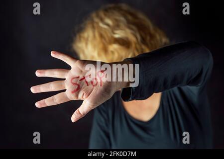 A blonde woman dressed in black covers her face with a hand on which she has written the word stop with a red marker, to stop violence against women Stock Photo