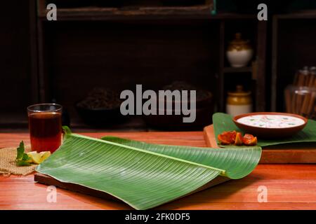 Banana leaf,fresh green banana leaf which is empty placed on wooden texture with a small bowl of raitha and a glass of lemon tea placed beside it, kit Stock Photo