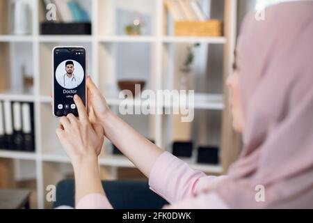 Close up blurred back view of young Muslim woman sitting at home, calling for online consultation with male Indian Arabian doctor on cell phone. Telemedicine and healthcare concept. Stock Photo