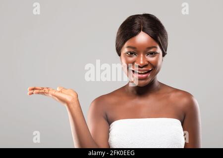 Pretty Black Female Wrapped In Towel After Bath Holding Something On Her Empty Palm, Demonstrating New Skincare Product, Beautiful African Woman Adver Stock Photo