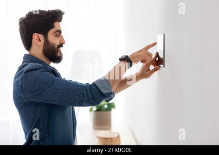 Smart home concept. Arab man using control system on digital tablet on wall, controlling house automation processes, side view, free space. Domestic I Stock Photo