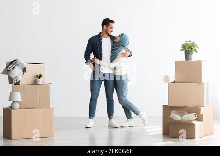 Loving muslim couple standing among moving supplies paper boxes with belongings in empty apartment, full length photo, copy space. Positive guy and yo Stock Photo