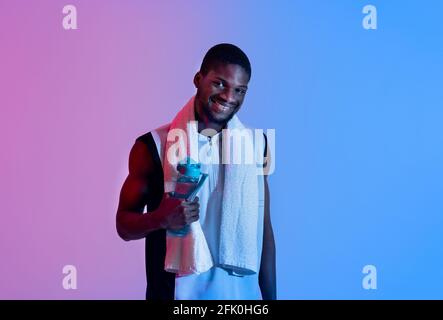 Young black sportsman with bath towel holding bottle of water in neon light. Positive African American athlete staying hydrated after workout or sauna Stock Photo
