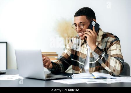 Satisfied smiling mixed race latino male freelancer or manager with glasses, have cell phone conversation, sits at work desk, uses laptop, checking e-mail, looking for information at internet Stock Photo