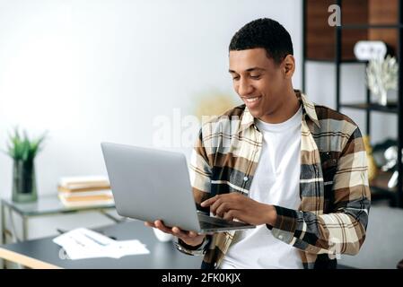 Smiling modern stylishly dressed mixed race latino freelancer guy, manager or student, stands near work desk, holds laptop, chatting online with friends, browses social networks, internet,checks email Stock Photo