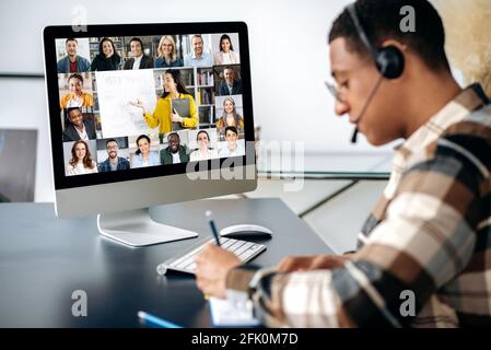 Successful mixed race clever male student learning distantly. At laptop screen female teacher and students. Teacher conducts online lecture, male student with headset takes notes. Video call Stock Photo