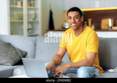 Headshot of friendly handsome young mixed race latino man, wearing orange t-shirt, sitting in living room on a couch with laptop, looking at camera with pleasant smile Stock Photo