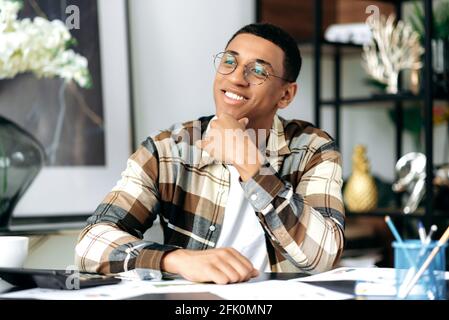 Portrait of a handsome contented mixed race hispanic young man, freelancer or student, wearing glasses and stylishly dressed, sitting at his work desk, looking away, smiling, thinking about weekend Stock Photo