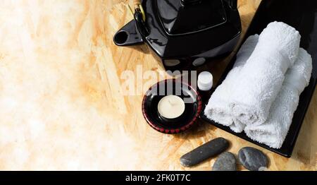 Black ceramic teapot in oriental style aroma lamp and aroma oil rolls of white towels on a ceramic dish and sea pebbles for face massage. Stock Photo