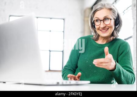 Smiling and friendly senior woman wearing headset and glasses using a laptop for video connect with colleagues, customers, a mature customer service representative takes a call, talking via video call Stock Photo