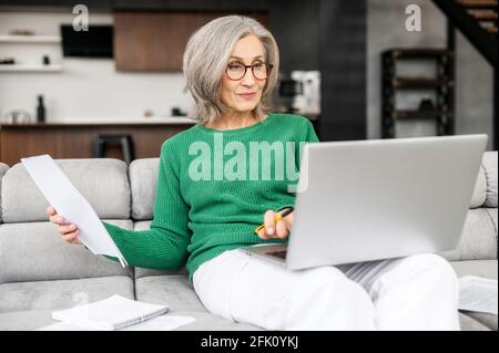 Confident retired woman is calculating house bills, checking spending per month, monitoring savings, senior female sits on the couch at home with a laptop and looking through workpapers, utility bills Stock Photo