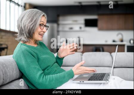 Middle-aged grey-haired woman talking online with family or friends, making video call on the laptop, senior female lying down on the sofa and enjoys video meeting on the computer Stock Photo