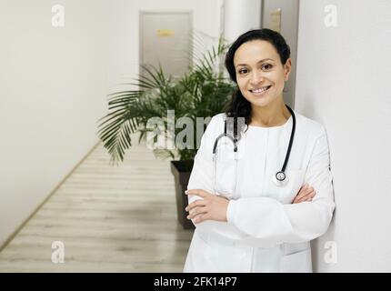 General practitioner. Positive female mixed-race doctor wearing a medical uniform with arms crossed at hospital corridor Stock Photo