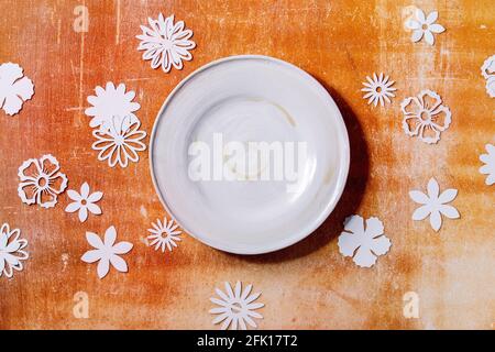 Empty white ceramic plate with paper flowers around over orange stone background. Flat lay, copy space Stock Photo