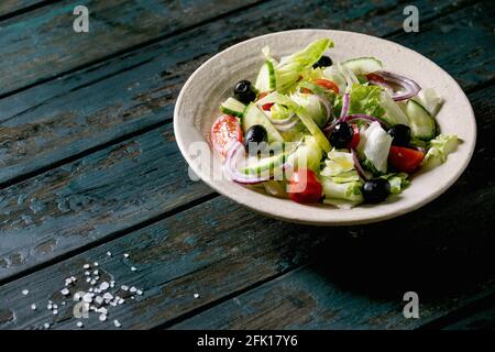 Classic vegetable salad with tomatoes, cucumber, onion, salad leaves and black olives in white ceramic plate. Dark wooden table. Stock Photo