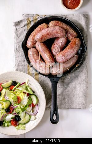Grilled italian sausages salsiccia in cast-iron pan served with tomato sauce and plate of fresh vegetable salad on white marble background. Balanced d Stock Photo