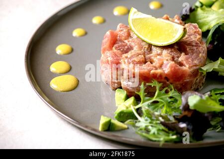 Tuna tartare with green salad, lime, avocado and mustard sauce serving on ceramic plate over white texture background. Fine dining, restaurant appetiz Stock Photo