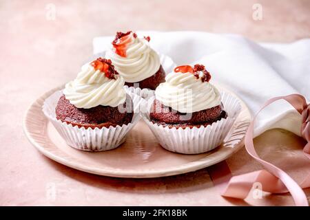 Homemade Red velvet cupcakes with whipped cream on pink ceramic plate, white napkin with ribbon over pink texture background. Stock Photo