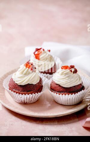 Homemade Red velvet cupcakes with whipped cream on pink ceramic plate, white napkin with ribbon over pink texture background. Stock Photo