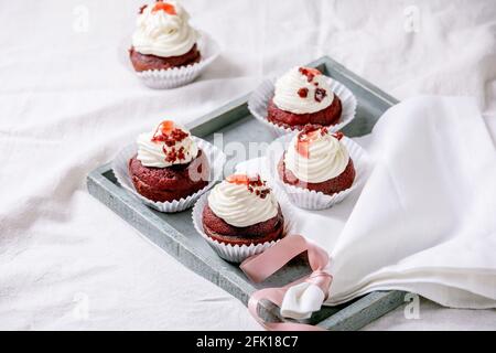 Homemade Red velvet cupcakes with whipped cream on wooden tray, white napkin with ribbon on white linen table cloth. Stock Photo