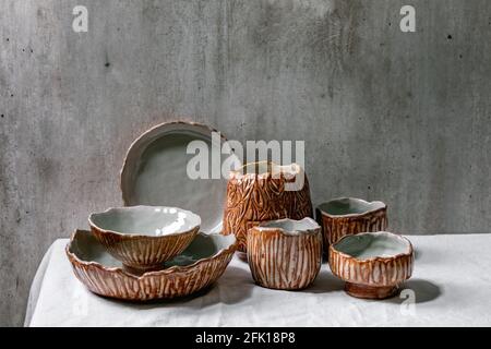 Set of empty craft ceramic bowls cover by grey and brown texture glaze on white linen tablecloth with gray wall behind. Copy space. Stock Photo