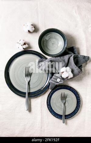 Set of empty gray ceramic plates with forks and textile napkin, decorated by cotton flowers over white linen tablecloth. Table setting. Flat lay, spac Stock Photo